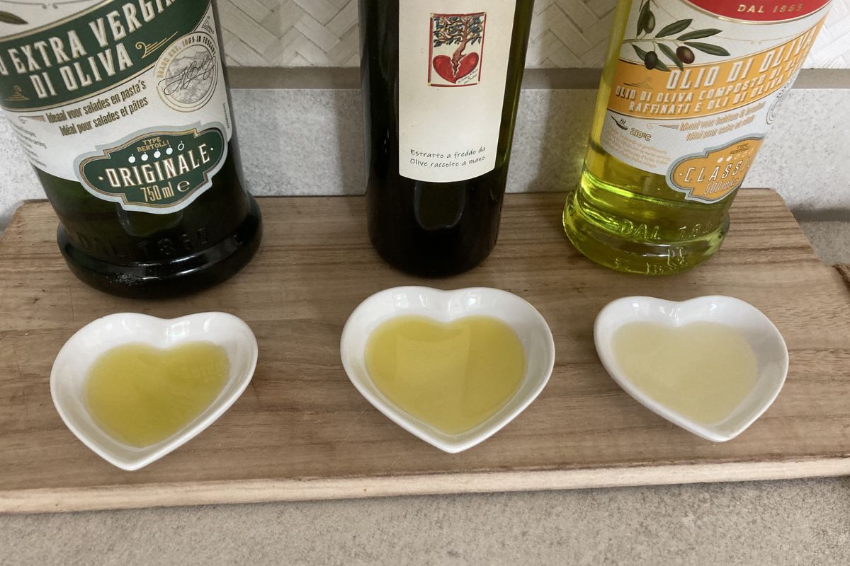Drie verschillende flessen Bertolli en Italiaanse olijfolie op een houten serveerplank, met daarvoor drie hartvormige schaaltjes gevuld met olijfolie voor een smaaktest tussen gewone en extra vierge olijfolie.